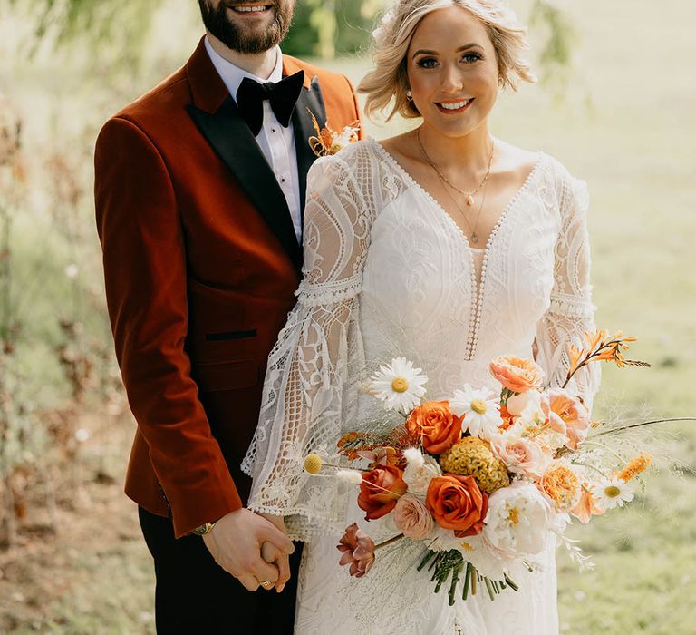 Groom in an orange tuxedo hugs the bride from behind in a boho lace wedding dress holding pretty bouquet 