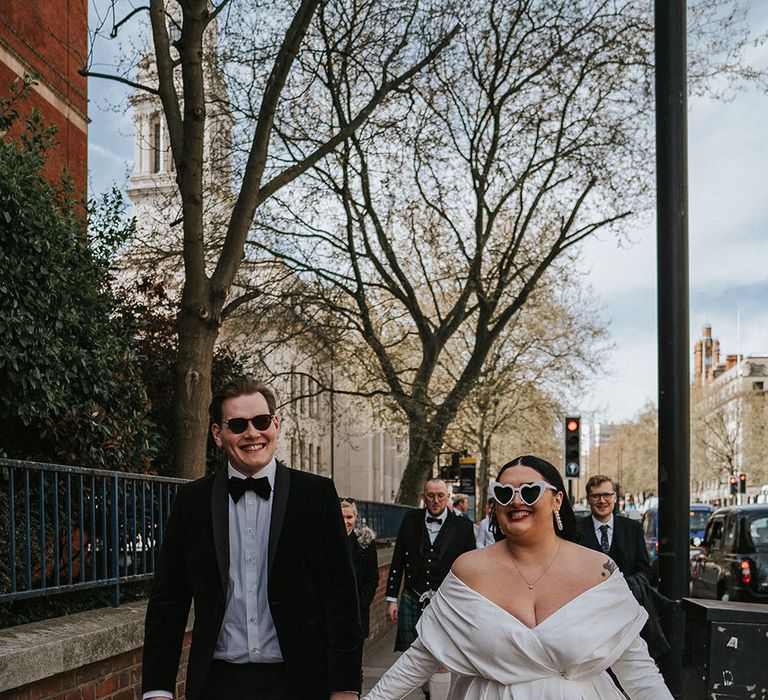 Bride wears off-the-shoulder ASOS curve wedding dress and walks hand in hand with her groom in black-tie through the city