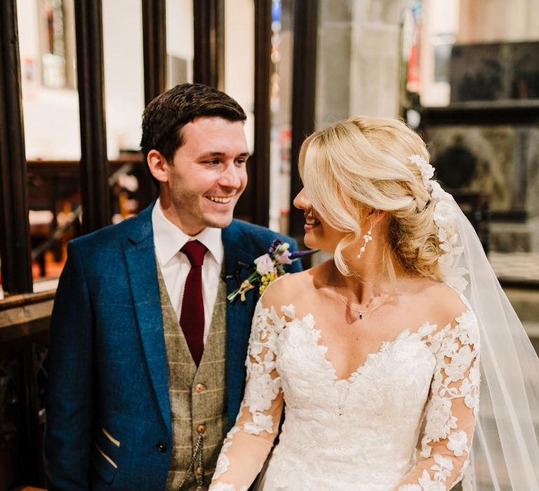 Groom wears three piece suit with blue jacket and maroon tie stood beside his bride wearing lace Pronovias wedding dress during church ceremony