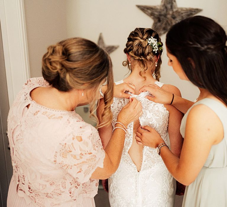 Bridesmaid in a soft green dress helps the mother of the bride get the bride into her lace wedding dress with button back detail 