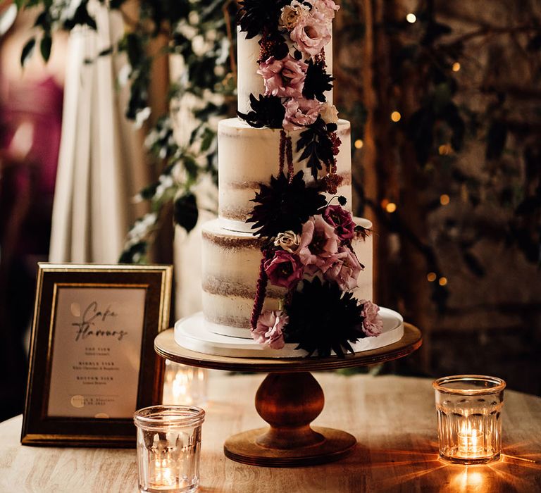 Three tier semi-naked wedding cake decorated with dark flowers on a wooden cake stand for the rustic wedding 