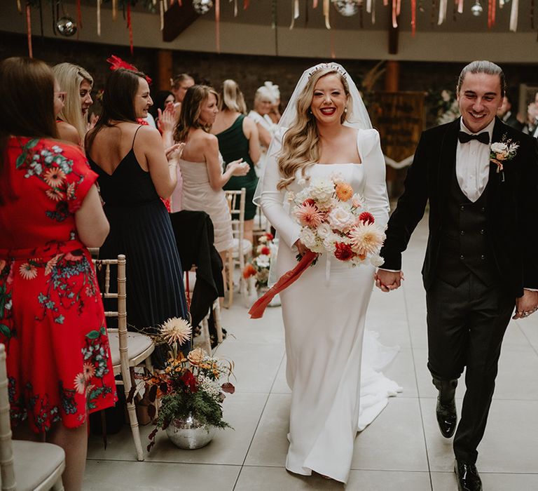 Bride in square neck satin wedding dress with two tier veil and colourful mixed floral bouquet with groom in black suit with floral boutonniere beginning confetti walk