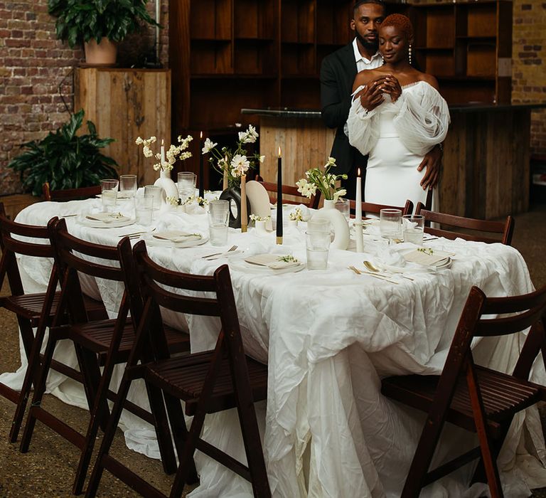 Bride in skinny black suit and bride in off shoulder wedding dress with lace puff sleeves standing by monochrome wedding table plan 