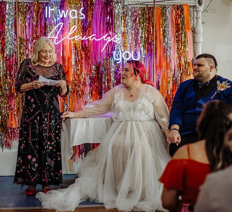 Bride in corset top wedding dress with leaf detailed lace overlay holding hands with groom in midnight blue velvet tuxedo and autumnal coloured boutonniere watching mother of the bride in black and floral overlay dress doing a speech 