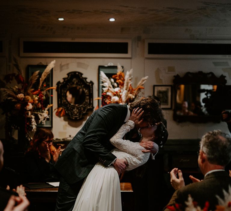 Bride in long sleeve lace vintage wedding dress and closed toe satin heels kissing groom in dark suit with orange tie at the alter of Autumnal wedding