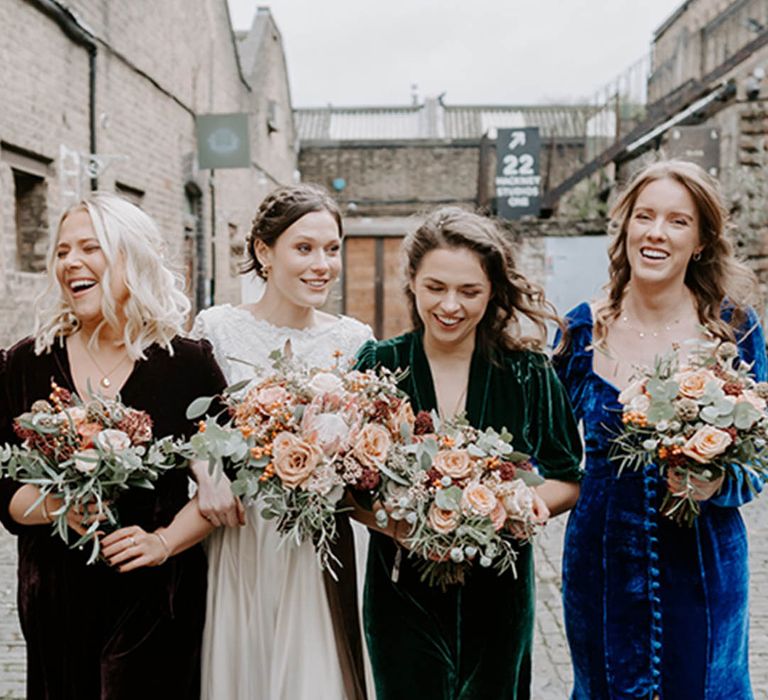 The bride in a lace wedding dress smiles as she walks with her bridesmaids in velvet dresses in different colours