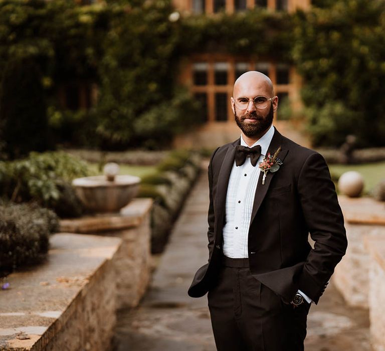 Groom in the garden of Oxfordshire wedding venue wearing classic black tuxedo, autumnal boutonniere and bowtie