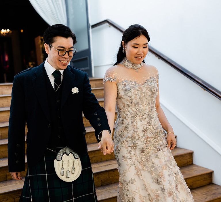 Bride in embellished bespoke reception dress walks down the stairs alongside her groom in green tartan kilt