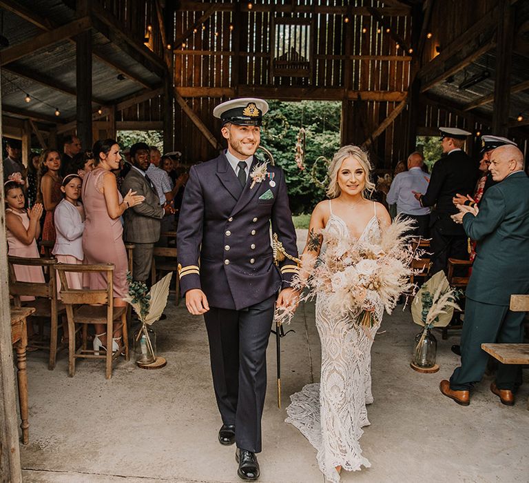 Groom in a double breasted military uniform with white hat and sword walking out of Nancarrow Farm barn wedding venue with the bride in a boho dress