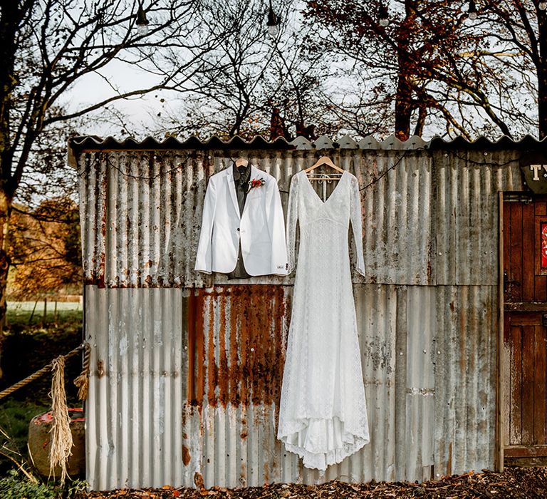 Long sleeve boho lace wedding dress and white grooms blazer hanging up on shed at Kent barn wedding venue UK 