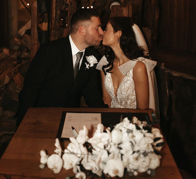 Groom in black suit and patterned tie wearing a white rose buttonhole kissing bride in sparkly wedding dress as they sign the wedding register 