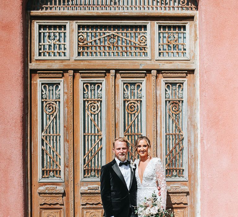 Bride in 3D lace wedding dress with long sleeves and shoulder pads standing with groom in black tuxedo with their precious pet dog 