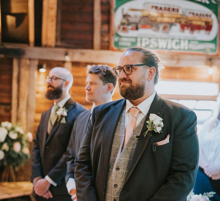 The groom wearing glasses in a dark grey suit with light grey checkered waistcoat stands at the altar for the wedding ceremony 