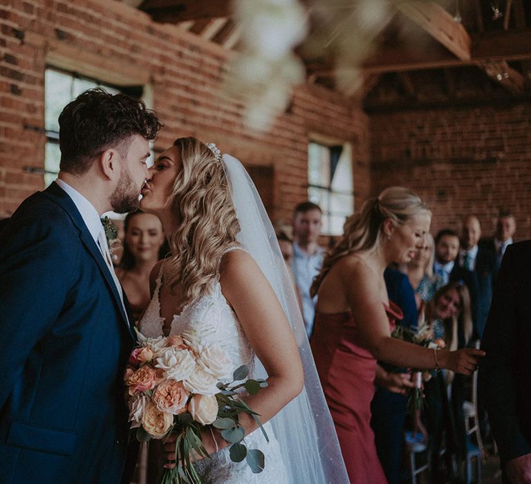 The groom in navy suit shares the first kiss as a married couple with the bride in a lace wedding dress with tiara and veil holding a white and peach rose bouquet