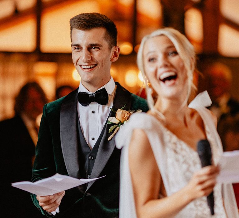 The groom smiles brightly in his velvet tuxedo whilst the bride laughs holding a microphone and speech during the wedding reception at Silchester Farm