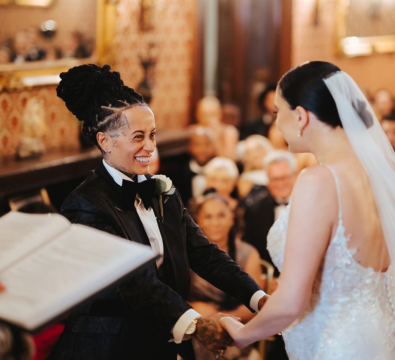 Bride in black snakeskin print tuxedo with black bowtie and white rose boutonniere holding hands with bride in lace sleeveless v neck Enzoani wedding dress with tulle finish and church length veil at Grittleton House