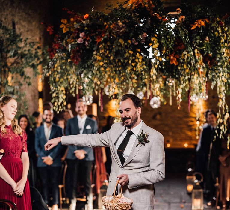 A flower man in a light grey suit walking down the aisle holding a wicker basket of flower petals 