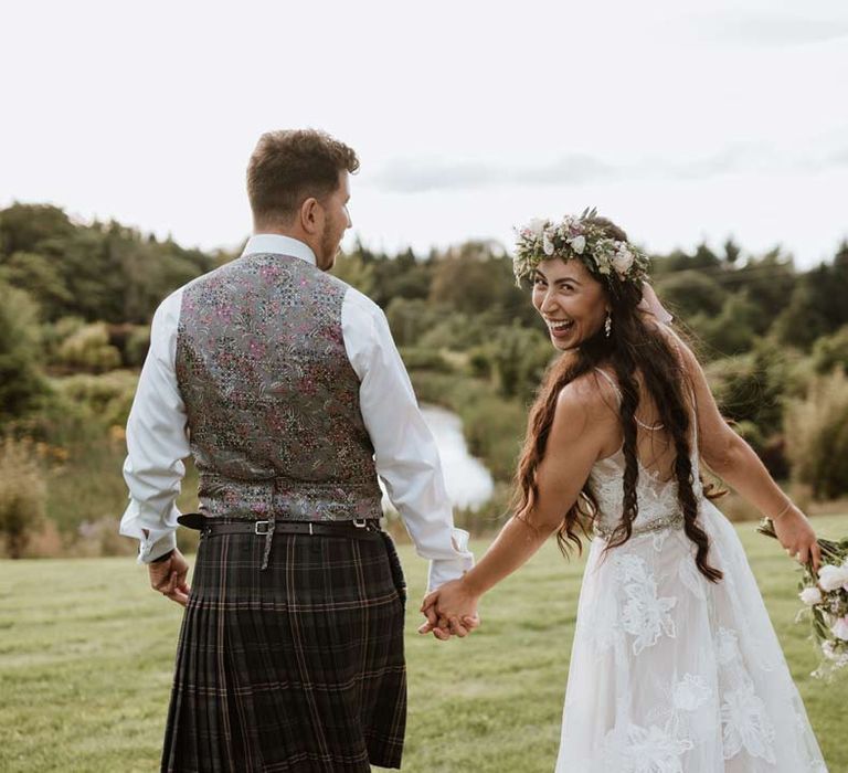 Bride in floral 3d applique wedding dress and white garden rose flower crown looking back at camera with groom in grey waistcoat and kilt 