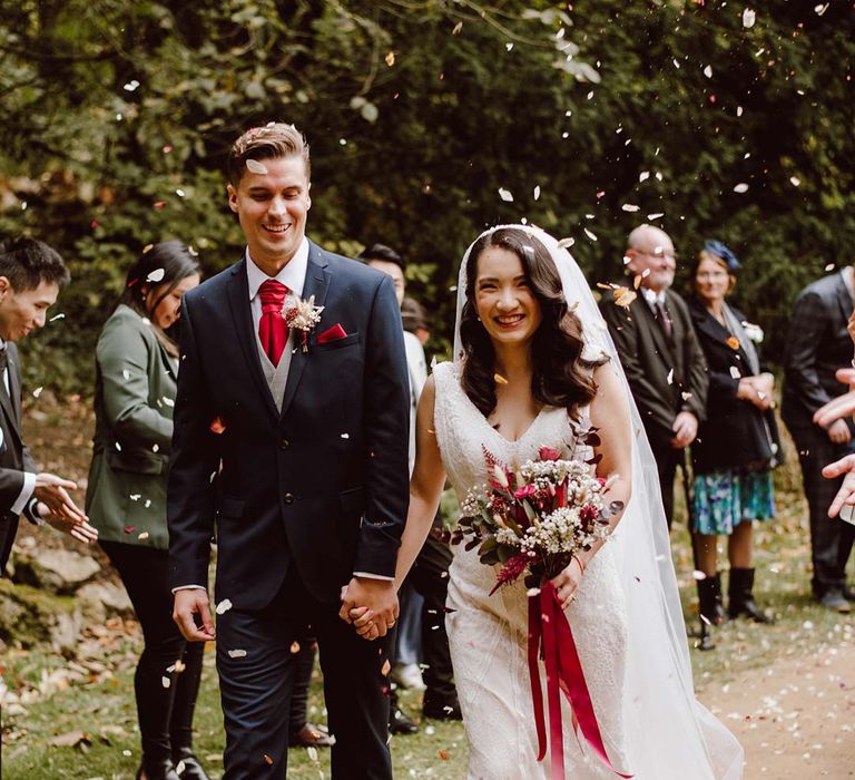 Bride in sleeveless lace wedding dress and church length red ombre veil holding large red rose and dried flower bouquet with red lace walking with groom in deep blue grooms blazer, grey waistcoat, red tie, red pocket square and mixed dried flower boutonniere doing wedding confetti exit 