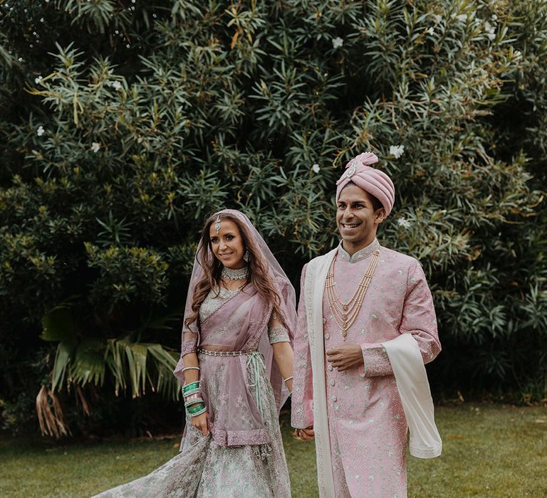 Groom in light pink sherwani, light pink turban and cream trousers walking with bride in mint green and dusky pink ombre embellished lehenga 