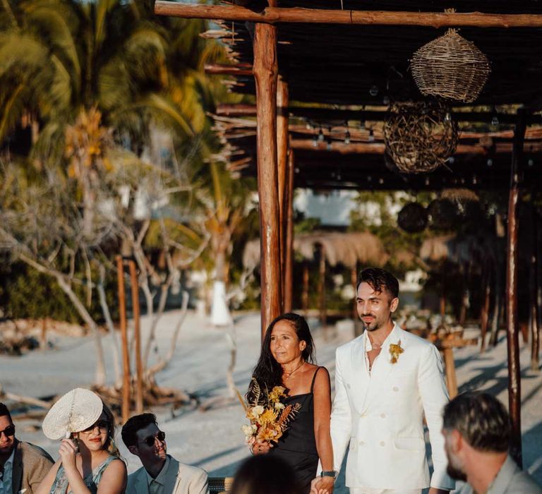 Groom walking down the aisle in white mens wedding suit, dusky orange garden rose and dried flower boutonniere, white shirt, and silver jewellery with wedding guest in black satin strappy dress 