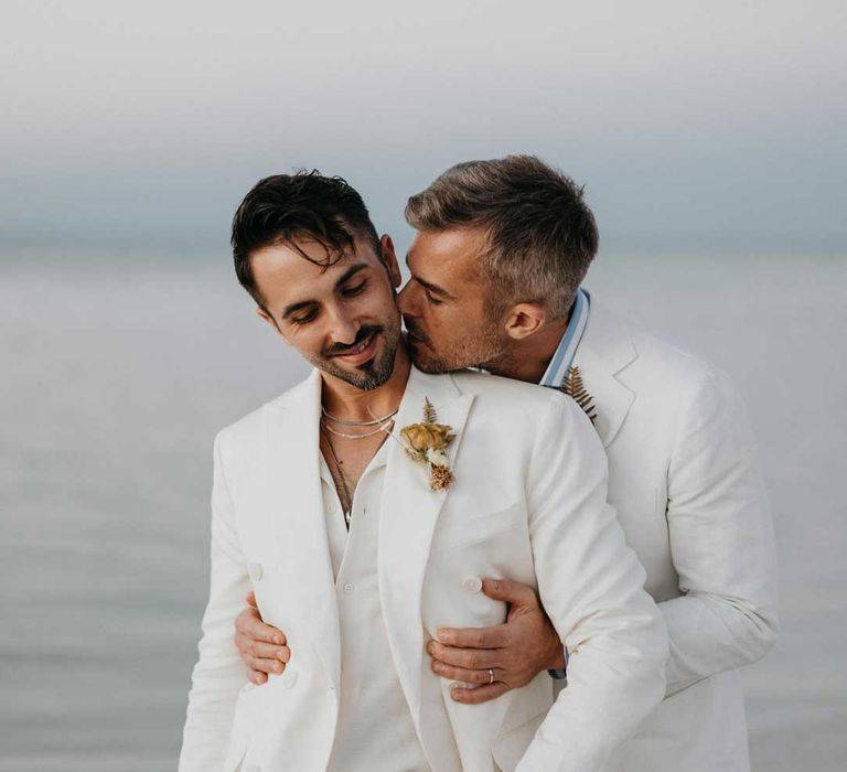 Groom in mens white wedding suit with dusky orange garden rose and dried flower boutonniere, white shirt and silver jewellery standing on the beach with Groom wearing white blazer, light blue and white striped shirt and dusky orange garden rose and dried flower boutonniere