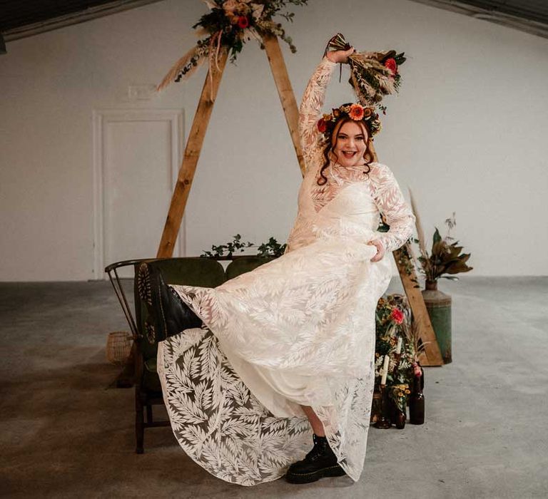 Bride in boho long sleeve 3D applique wedding dress with dried flower crown holdingearthy toned garden rose, pampas grass, dried flower and foliage bridal bouquet sitting on crushed velvet olive green loveseat in front of one small and one large wooden triangular wedding arch with pampas grass, foliage, and garden rose arrangements attached, rustic wedding decorations, large plants in vases and pillar candles 