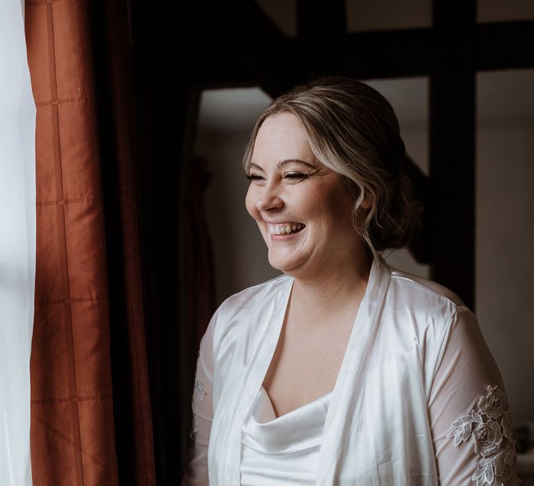 Bride in satin nightgown and robe looking out the window as she gets ready for the wedding day 