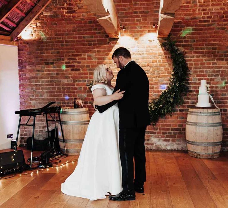 Groom in classic black grooms three piece tuxedo with white rose boutonniere and black bowtie doing wedding first dance with bride in satin off the shoulder wedding dress at Brickhouse Vineyard wedding venue 