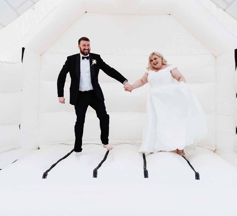 Bride in satin off the shoulder wedding dress holding hands with groom wearing classic black grooms three piece tuxedo with white rose boutonniere and black bowtie jumping on white wedding bouncy castle