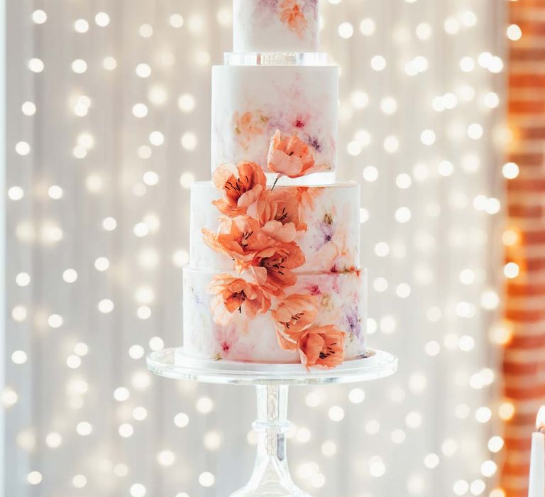 Four tiered hand-painted iced wedding cake with hand-made edible flower decorations on clear cake stand at Rackleys Barn with wedding fairy lights curtain