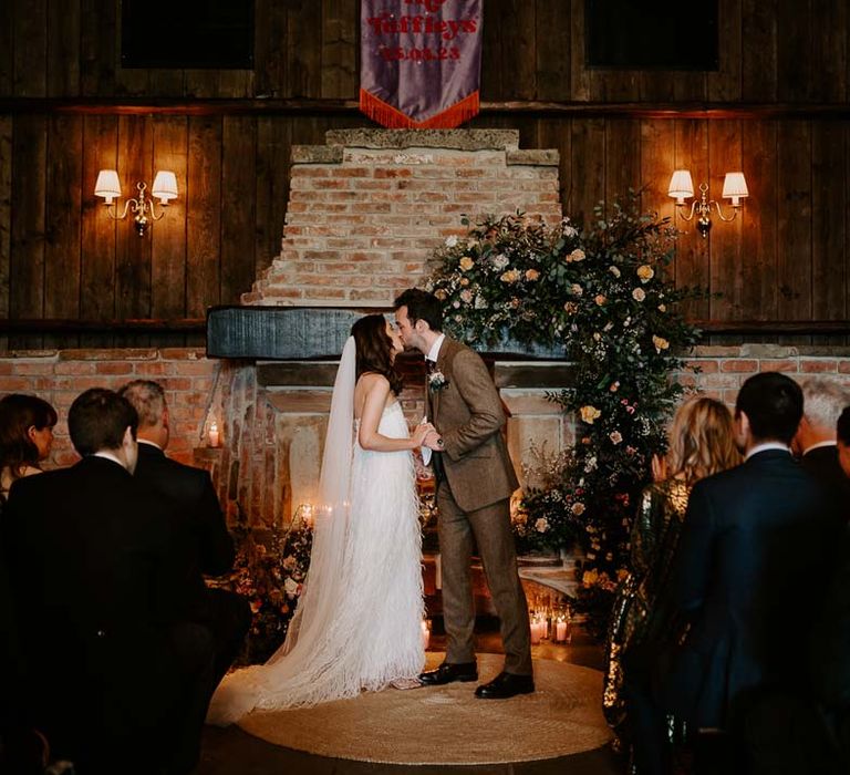 Groom in chocolate brown grooms suit kissing bride in strapless ivory fringed crepe gown and chapel length veil at the alter of Willow Marsh Farm Loughborough 