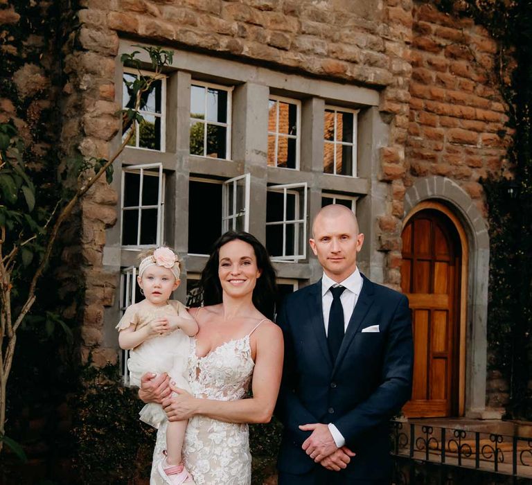 Groom in classic black grooms suit with white pocket square standing with bride in lace sleeveless wedding dress with puddle train holding baby in off-white dress and sequin headband with pink carnation