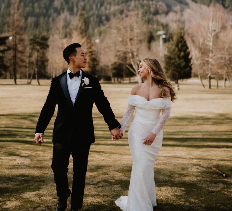 Groom in classic black tuxedo, black bowtie and white garden rose boutonniere walking hand in hand with bride in off shoulder sparkly mermaid wedding dress at French destination ski wedding