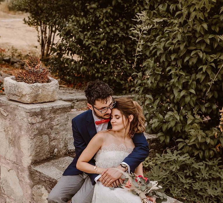 groom in grey chinos navy blazer and orange bow tie embracing his boho bride in a strapless lace wedding dress at greece destination wedding