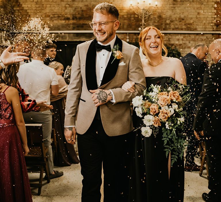 Groom in beige and black tuxedo walking with bride in black bridal gown carrying orange rose cascading bouquet 