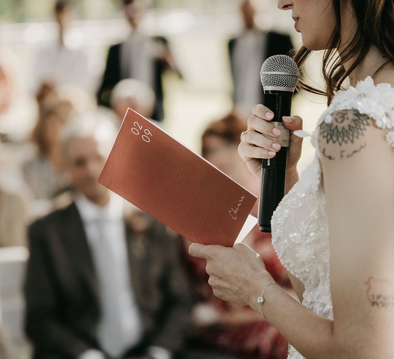 bride reading her wedding vows from an orange vow booklet at outdoor boho wedding in Italy