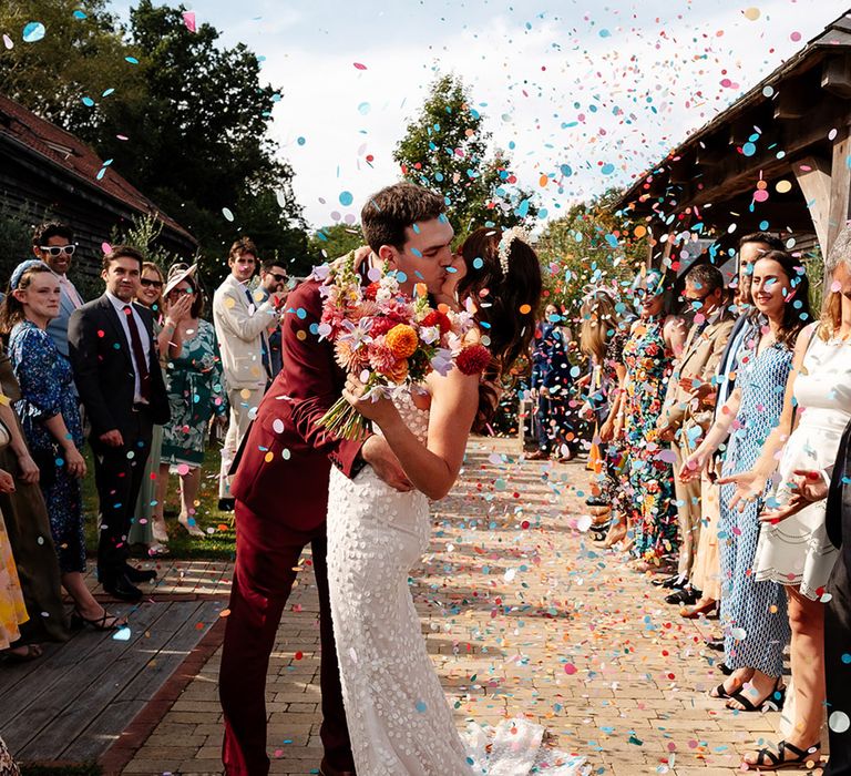 The bride and groom kiss as guests throw colourful confetti over them in celebration 