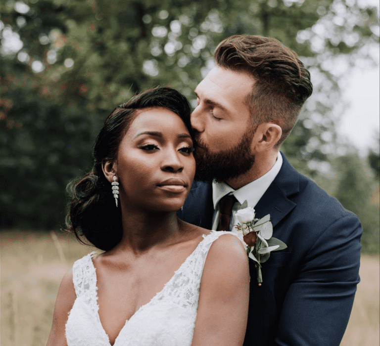 A couple embrace for couples photo at their wedding. The bride wears a v neck dress and has a low bun in her hair.