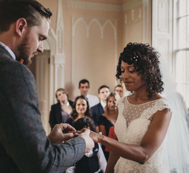 A couple stand at the altar exchanging rings. The bride has short curly hair and wears a veil as the groom puts a ring on her finger.