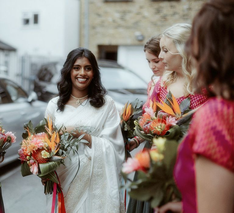 Asian bride in a white sari holding a colourful tropical flower wedding bouquet surrounded by her bridesmaids in pink and green lenghas