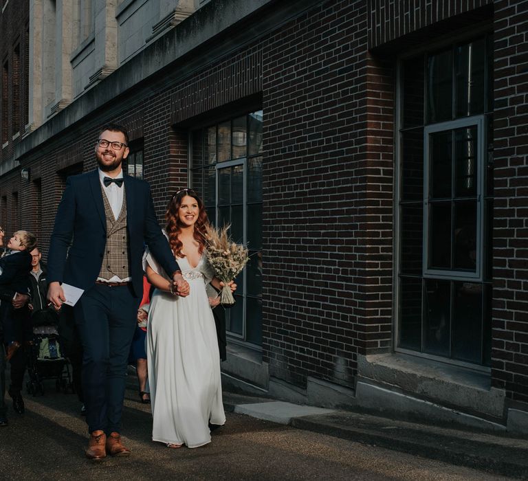 Bride holding dried flower bouquet and groom in navy suit with tartan waistcoat walk along the street 