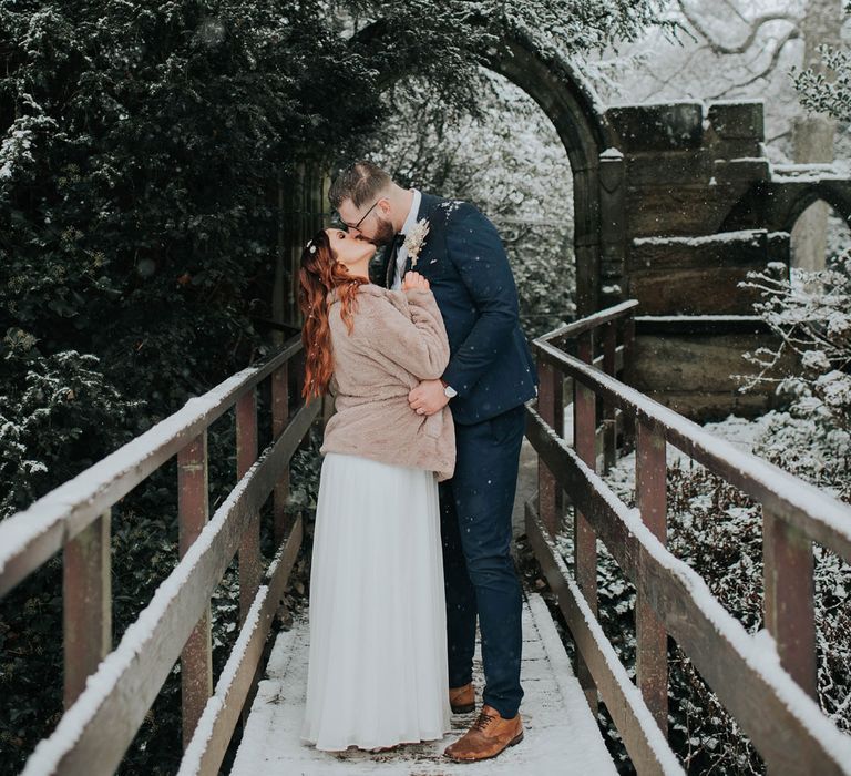 Bride in fur coat stands on bridge with groom in navy suit during snowfall at Cannon Hall