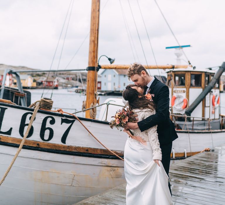 The bride and groom kiss next to a white fishing boat at their destination wedding in Sweden