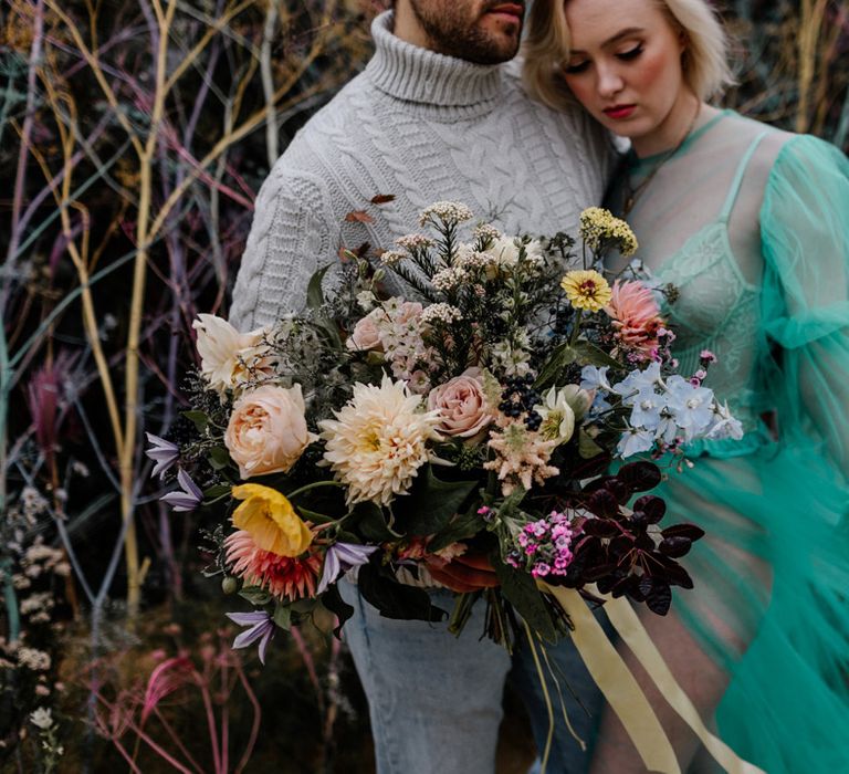 Groom in a grey cable jumper holding a colourful flower wedding bouquet tied with ribbon 