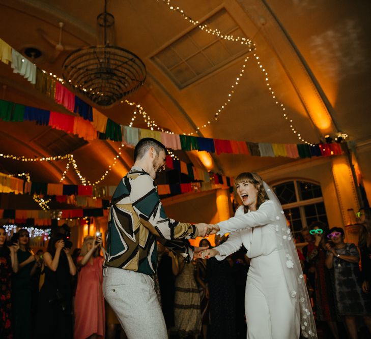 Groom in grey trousers and a grey and mustard pattern shirt dancing with his bride in a jumpsuit and appliqué veil 