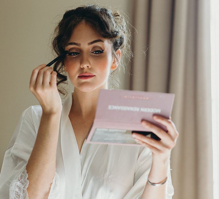 Bride on the wedding morning in a white getting ready robe putting on her own makeup 