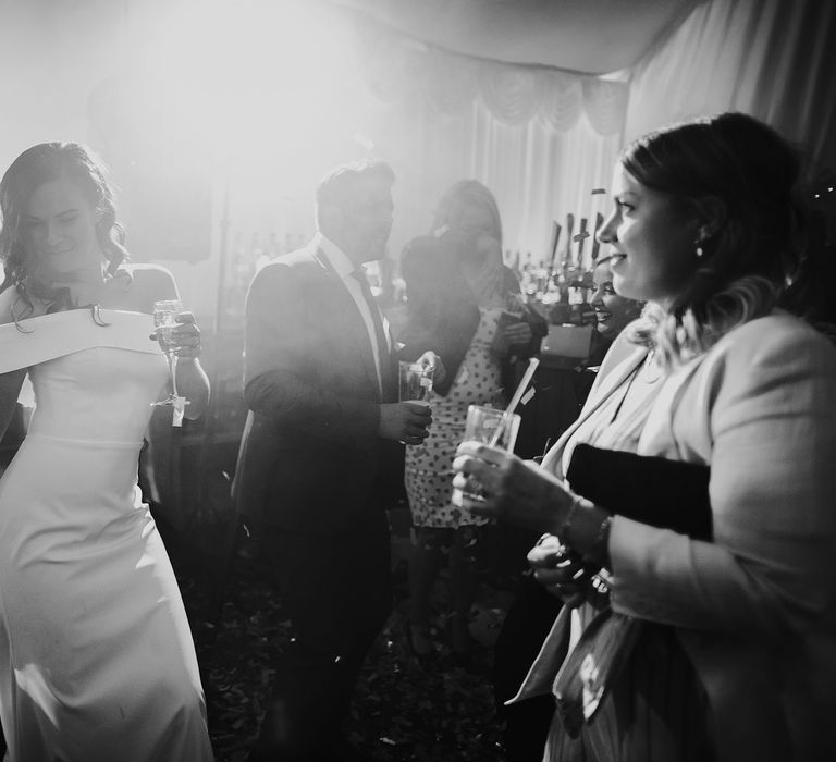 Bride in white strapless Vagabond wedding dress holding train and glass of sparkling wine dances to wedding band in marquee at lakeside wedding