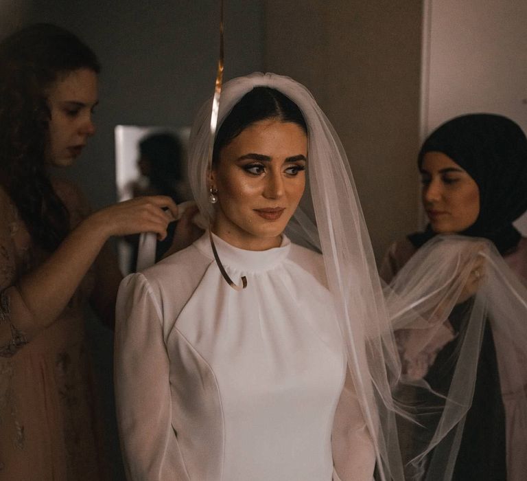 Black haired bride stands as wedding party help her get ready on the morning of her wedding