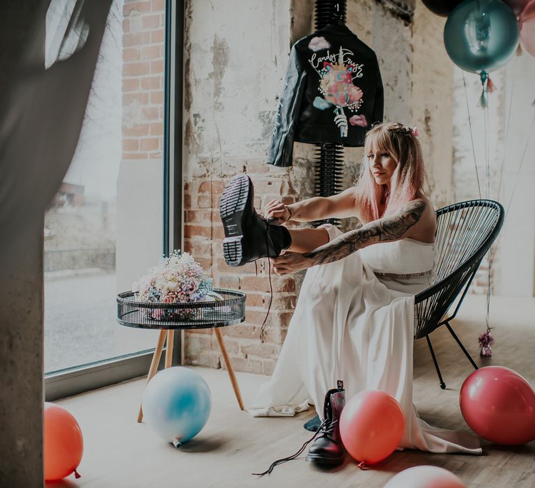 Bride in separates putting on her wedding boots whilst sitting on a black wire chair with her pastel bouquet and personalised leather jacket close by